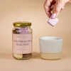 Jar of marshmallows selection with a hand holding a marshmallow over a ceramic cup on a beige background