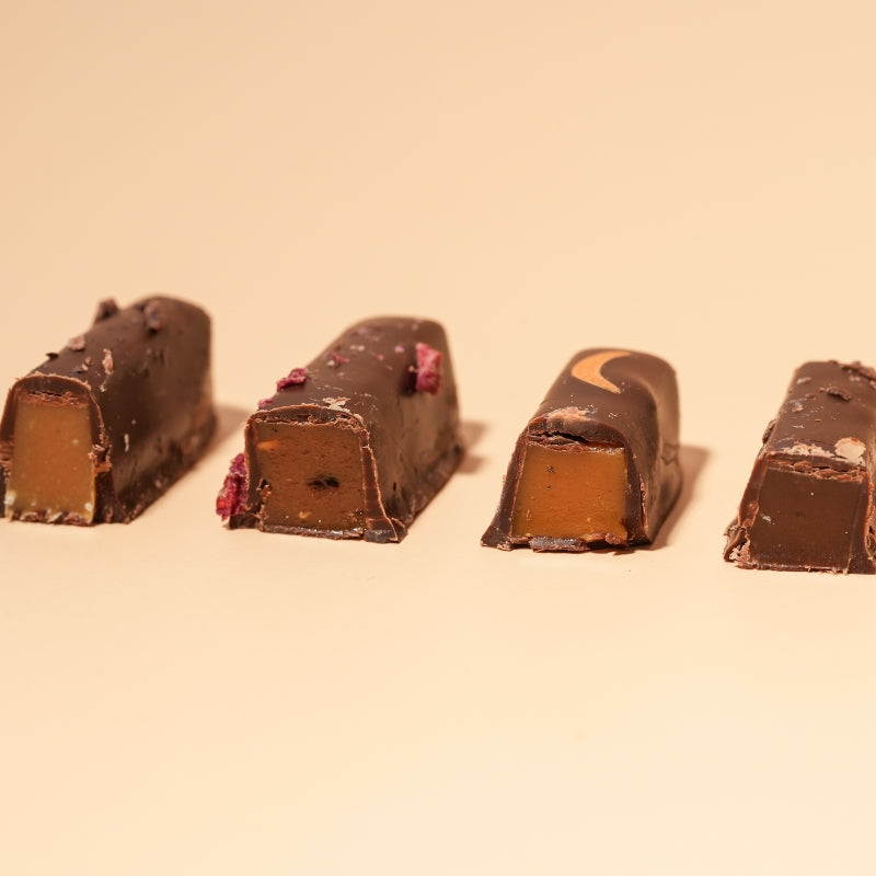 Four chocolate-covered caramel bars arranged in a row on a plain beige background.