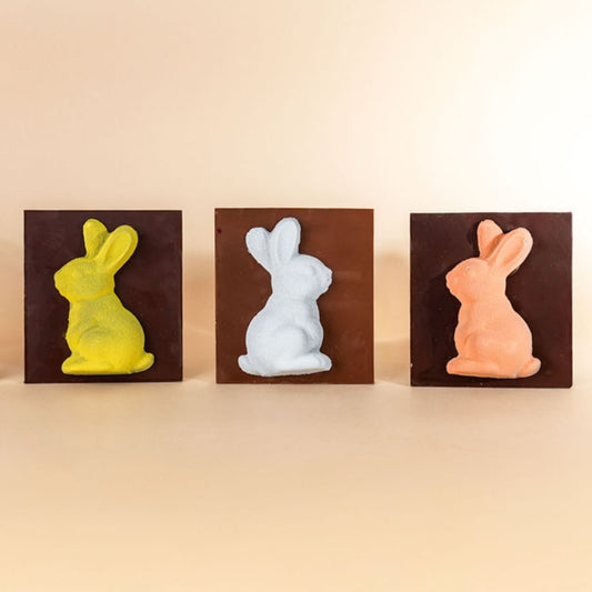 Three chocolate bunny molds in yellow, white, and orange on a beige background