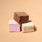 Three square marshmallows stacked in a pyramid on a warm peach background: one dark brown, one pink, and one off-white