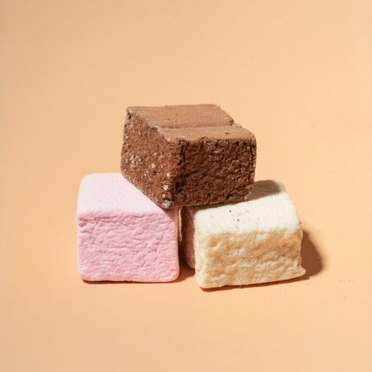 Three square marshmallows stacked in a pyramid on a warm peach background: one dark brown, one pink, and one off-white