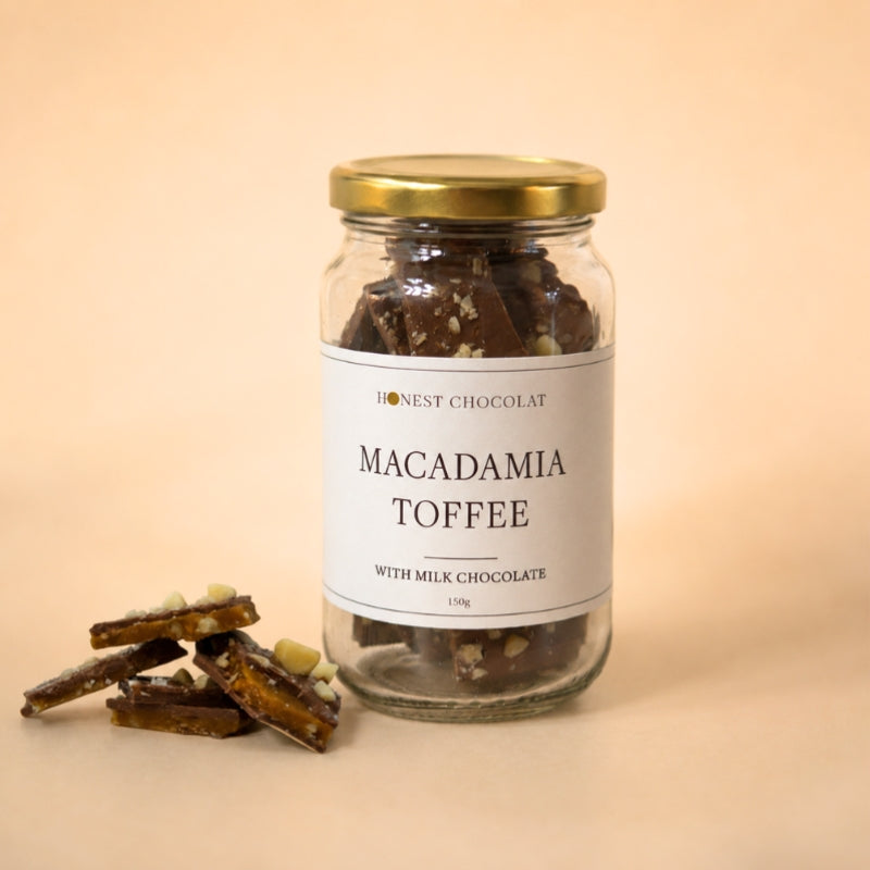 A glass jar of Honest Chocolat macadamia butter toffee with milk chocolate, featuring a white label and a gold lid, set against a warm beige background with a few pieces of the toffee displayed in the foreground.