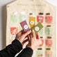 A person's hands hold three individually wrapped chocolate bars in front of a large, blurred advent calendar. The chocolate bars are wrapped and have round white labels with handwritten text showing the chocolate flavours.