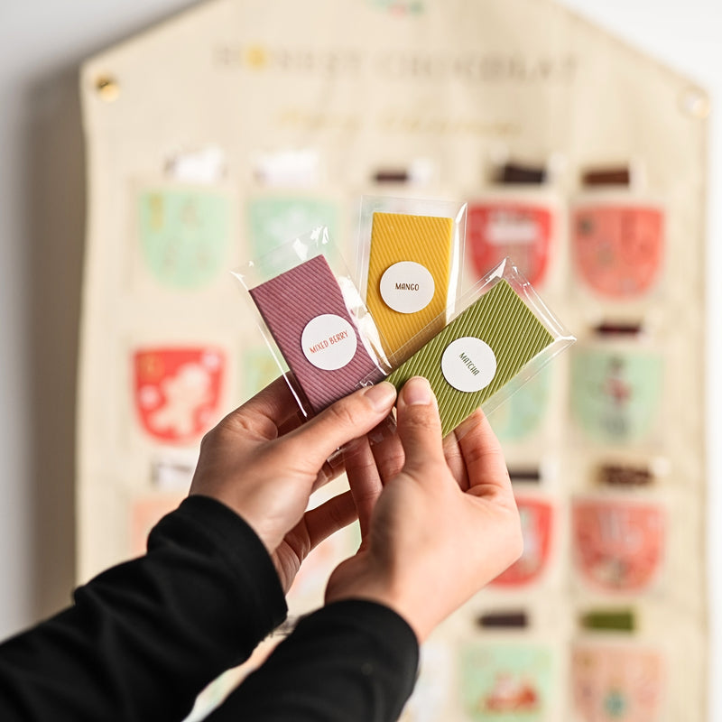 A person's hands hold three individually wrapped chocolate bars in front of a large, blurred advent calendar. The chocolate bars are wrapped and have round white labels with handwritten text showing the chocolate flavours.
