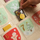 A close-up of a Christmas advent calendar with fabric pockets, each decorated with embroidered holiday motifs and numbers. A hand is opening a pocket, which contains a wrapped chocolate bar labeled 'CRANBERRY APRICOT & PISTACHIO'.
