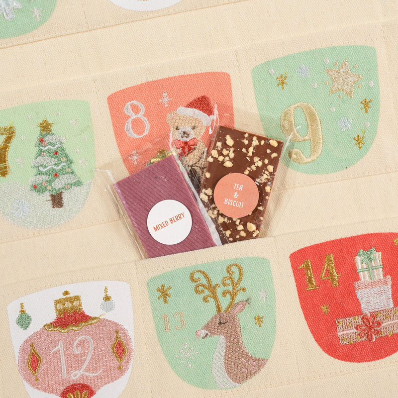 An embroidered Christmas advent calendar fabric panel with numbered sections and festive illustrations, showing two packaged treats placed in the center compartments. The flavours for the chocolate bars are Mixed Berry and Tea and Biscuit.