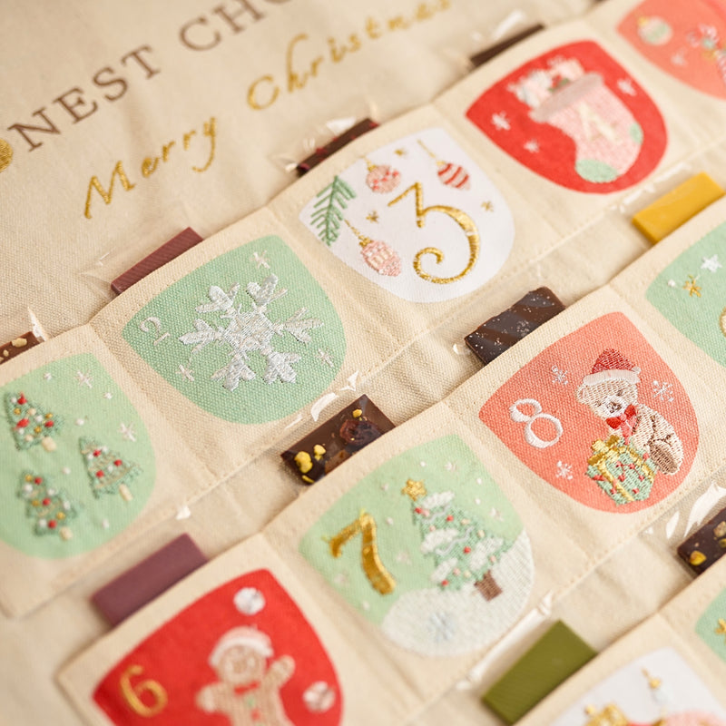 Close-up of a Christmas advent calendar with embroidered numbered pockets, festive illustrations, and small artisan chocolate treats inside the pockets of the tapestry calendar.